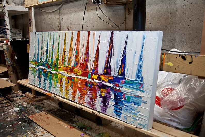 May include: A colorful abstract painting of sailboats on a white background. The painting is done in a thick impasto style, with visible brushstrokes. The sailboats are painted in a variety of bright colors, including red, orange, yellow, green, blue, and purple. The water is painted in a lighter blue and white, with hints of green and yellow. The painting is framed in a white frame.