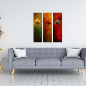 May include: Three abstract paintings of trees in different colors. The first painting is green with a yellow tree, the second is orange with a red tree, and the third is red with a green tree. The paintings are hanging on a white wall above a gray sofa.