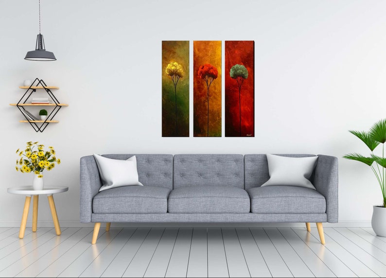 May include: Three abstract paintings of trees in different colors. The first painting is green with a yellow tree, the second is orange with a red tree, and the third is red with a green tree. The paintings are hanging on a white wall above a gray sofa.