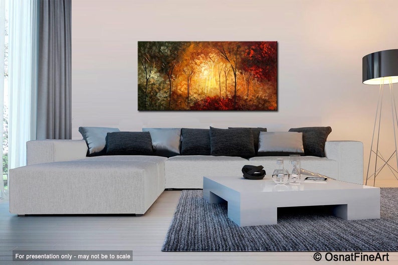 May include: Abstract painting of a forest scene with trees in shades of green, yellow, orange, and red. The painting is displayed on a white wall above a white sectional sofa with gray pillows and a white coffee table.