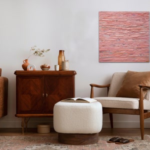 May include: A living room interior with a brown leather sofa, a white ottoman, a wooden armchair, a wooden cabinet, and a black metal stand with a black rotary phone. A pink abstract painting hangs on the wall.