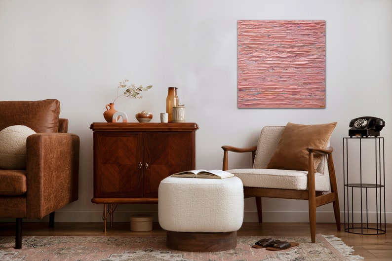 May include: A living room interior with a brown leather sofa, a white ottoman, a wooden armchair, a wooden cabinet, and a black metal stand with a black rotary phone. A pink abstract painting hangs on the wall.