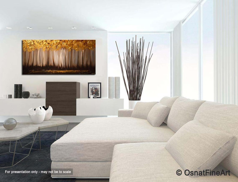 May include: Abstract painting of a forest with gold and brown tones. The painting is hanging on a white wall in a modern living room with a white couch and pillows.