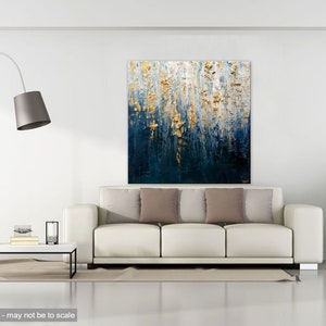 May include: Abstract wall art with a textured design in shades of blue, white, and gold. The artwork hangs above a white sofa with neutral-colored pillows. The room is minimalist with a modern floor lamp and a potted plant.