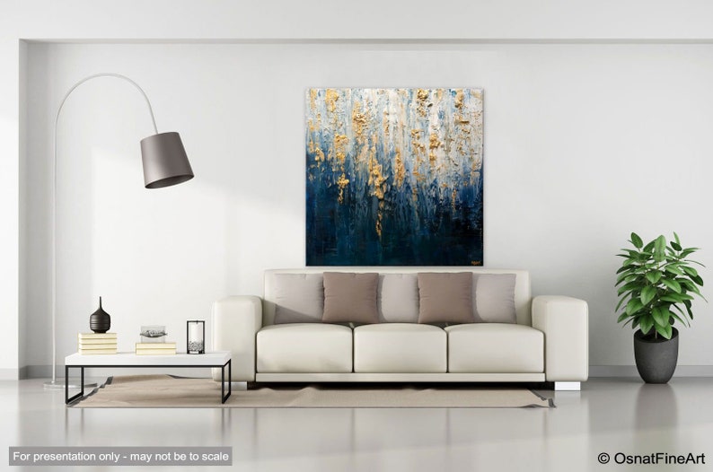 May include: Abstract wall art with a textured design in shades of blue, white, and gold. The artwork hangs above a white sofa with neutral-colored pillows. The room is minimalist with a modern floor lamp and a potted plant.