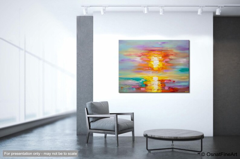 May include: Abstract painting of a colorful sunset over water. The painting is done in a palette of pink, orange, yellow, and blue. The painting is displayed on a white wall in a modern interior.