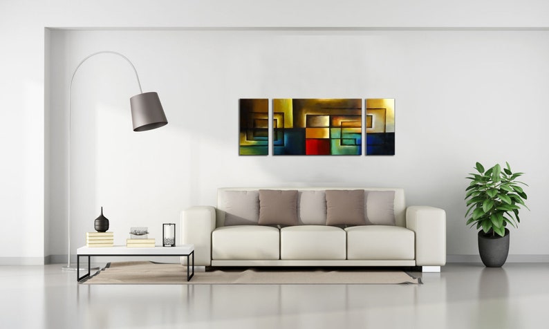 May include: Abstract art painting with a geometric design in shades of brown, blue, green, red, and yellow. The painting is divided into three panels and is displayed on a white wall above a white couch.