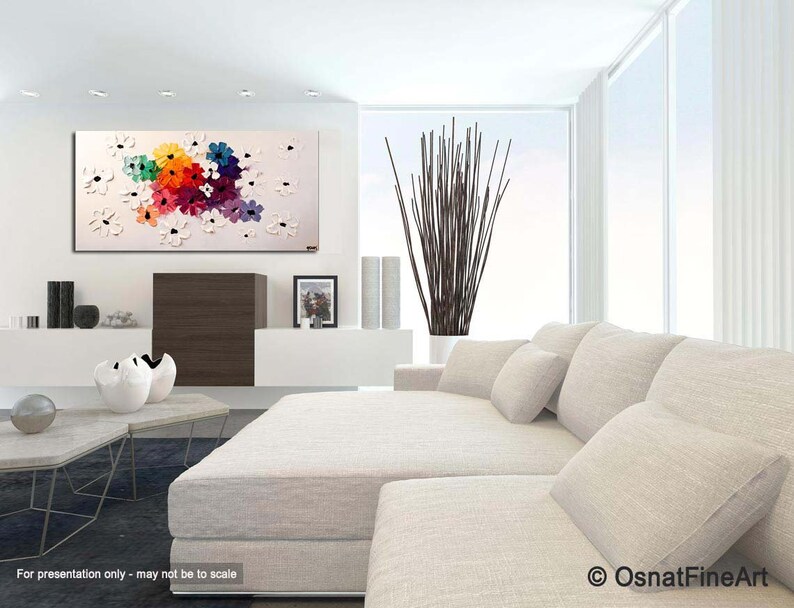 May include: Abstract painting of colorful flowers on a white background. The painting is displayed in a modern living room with a white couch and a coffee table.