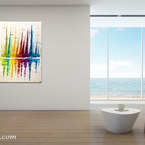May include: Abstract painting of colorful sailboats reflected in the water. The painting is on a white canvas and is framed in a white frame. The painting is hanging on a white wall in a modern living room with a large window overlooking the ocean.