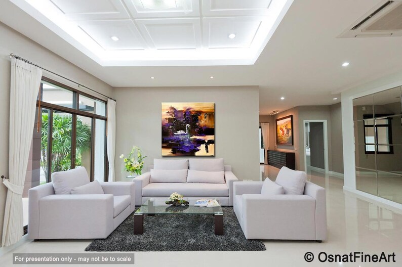 May include: Abstract painting of a white swan on a purple lake with a gold and brown background. The painting is hanging on a wall in a living room with white couches and a coffee table.
