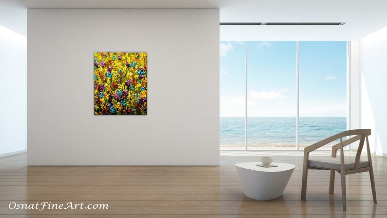May include: Abstract painting of a field of yellow, blue, and purple flowers. The painting is displayed on a white wall in a modern living room with a view of the ocean.