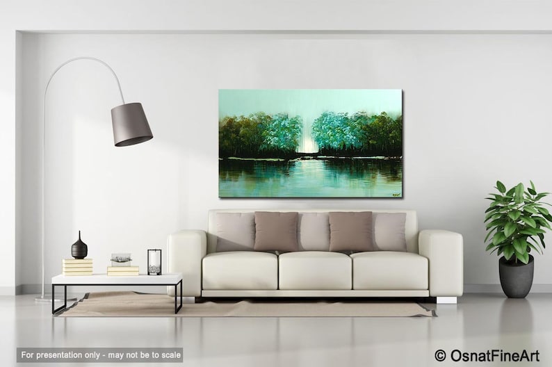 May include: A painting of a lake scene with trees on either side of the water. The trees are a mix of green and blue hues, and the water is a light blue with reflections of the trees. The painting is framed and hanging on a white wall in a living room setting.