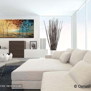 May include: Abstract painting of trees in a landscape with yellow, orange, brown, blue and green colors. The painting is hanging on a white wall in a living room setting with a white couch and a coffee table.