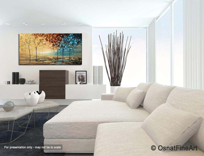 May include: Abstract painting of trees in a landscape with yellow, orange, brown, blue and green colors. The painting is hanging on a white wall in a living room setting with a white couch and a coffee table.