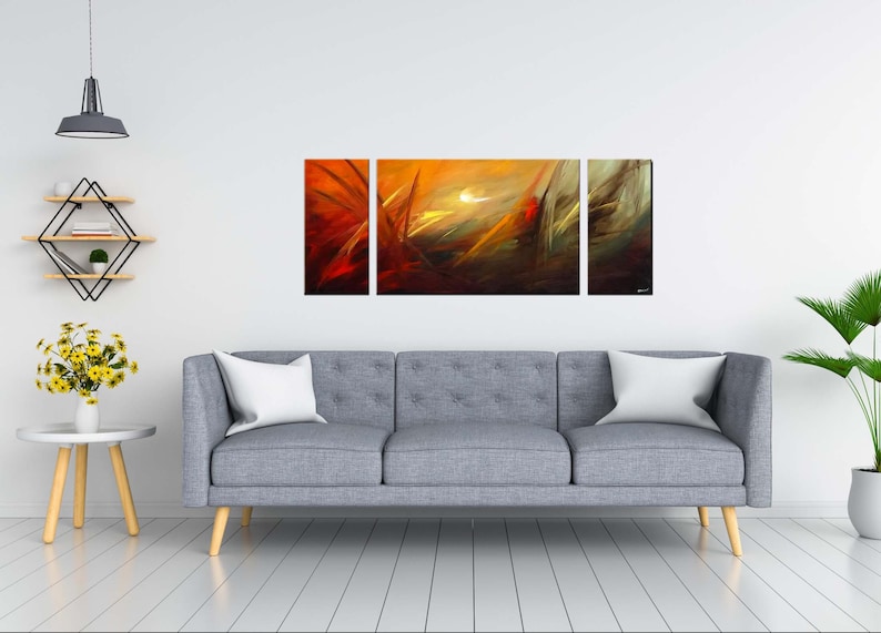 May include: A three-piece abstract art print set featuring a vibrant orange, yellow, and red color scheme. The artwork depicts a dynamic, swirling pattern with a hint of white in the center. The prints are displayed on a gray sofa in a modern living room setting.