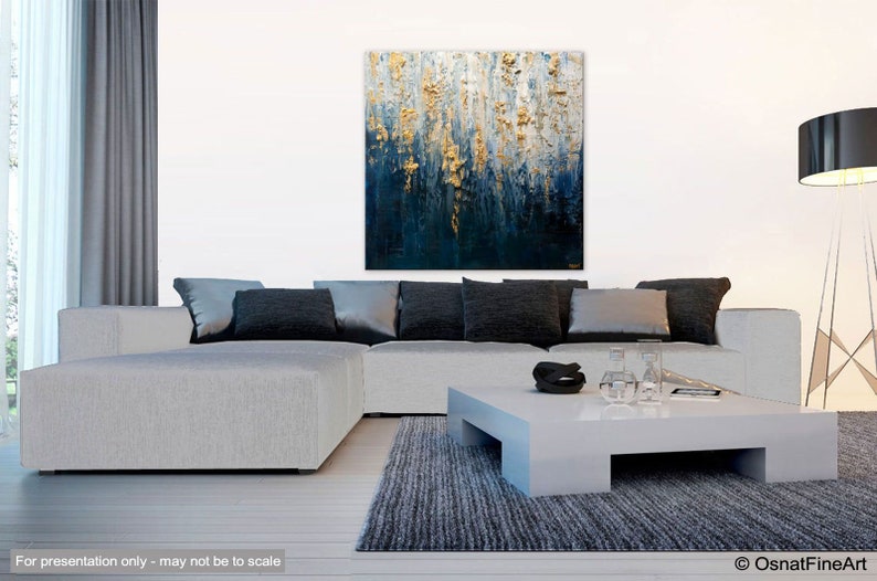 May include: Abstract wall art featuring a textured painting with gold and blue tones. The artwork hangs above a light gray sectional sofa with dark gray and silver pillows. A white coffee table and a gray rug complete the living room setting.