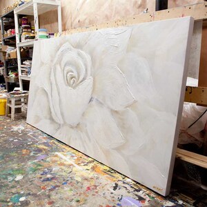 May include: A large white canvas painting of a white rose with textured brushstrokes. The painting is leaning against a wooden easel in an art studio.
