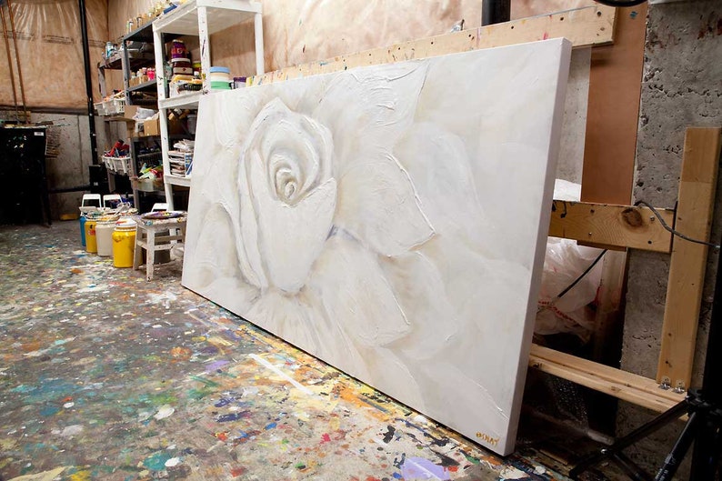 May include: A large white canvas painting of a white rose with textured brushstrokes. The painting is leaning against a wooden easel in an art studio.