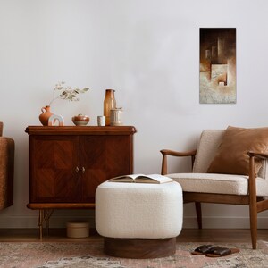 May include: A living room interior with a brown leather sofa, a white ottoman, a brown armchair, a wooden cabinet, and a black metal stand with a black rotary phone. A painting with brown and white geometric shapes hangs on the wall.