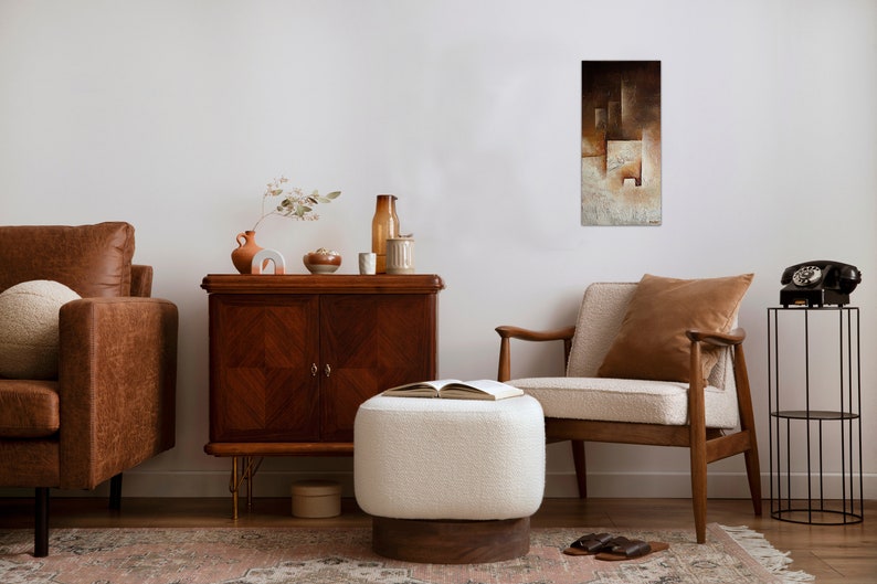 May include: A living room interior with a brown leather sofa, a white ottoman, a brown armchair, a wooden cabinet, and a black metal stand with a black rotary phone. A painting with brown and white geometric shapes hangs on the wall.
