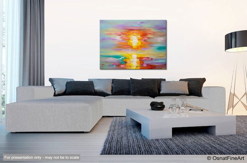 May include: Abstract painting of a colorful sunset over a body of water. The painting is done in shades of pink, orange, yellow, green, and blue. The painting is displayed on a white wall above a gray couch and a white coffee table.