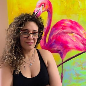 May include: A woman with curly hair wearing glasses and a black tank top stands in front of a painting of a pink flamingo on a yellow background.