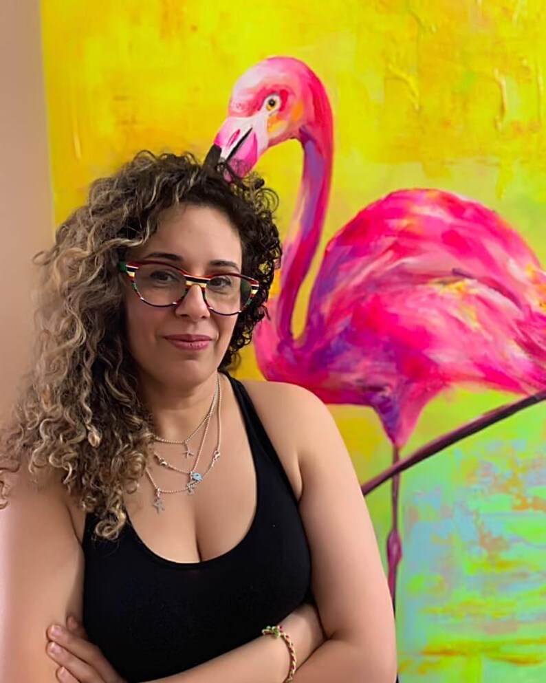 May include: A woman with curly hair wearing glasses and a black tank top stands in front of a painting of a pink flamingo on a yellow background.