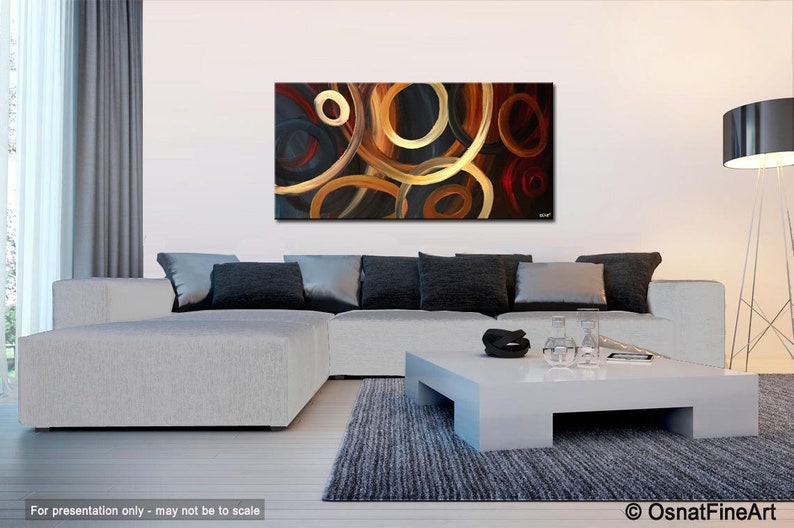 May include: Abstract painting with a black background and overlapping circles in shades of gold, brown, and red. The painting is displayed on a wall in a living room setting with a white couch and a coffee table.