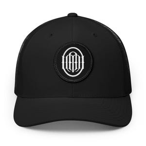 Puede incluir: Una gorra de camionero negra con un parche circular en el panel frontal. El parche muestra un logotipo de monograma bordado en blanco que consiste en letras estilizadas dentro de un borde ovalado. La gorra tiene un panel frontal sólido y malla.