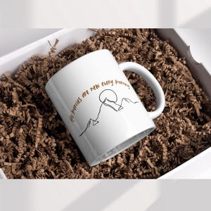 May include: White ceramic mug with a curved handle, featuring a black line drawing of a mountain range and sun. The mug has the text "His mercies are new every morning" in a warm brown color. The mug is presented in a white gift box with brown crinkle paper.