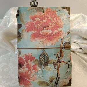Könnte beinhalten: Ein Notizbuch mit hellblauem Einband, großem rosa Blumenmuster, braunen Zweigen und goldenen Metallecken. Ein dünnes braunes Gummiband hält einen hängenden bronzefarbenen Blattanhänger, und ein eulenartiges Lesezeichen ragt oben heraus.