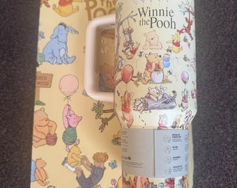 May include: A cream-coloured stainless steel tumbler featuring a Winnie the Pooh graphic print with various characters, a white handle, and a clear lid. The tumbler is positioned in front of a matching rectangular box with the same character illustrations.