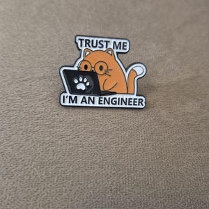 May include: An enamel pin featuring an orange cat wearing round glasses while working on a black laptop with a white paw print logo. The text above the cat reads TRUST ME and the text below the cat reads I'M AN ENGINEER in black capital letters.
