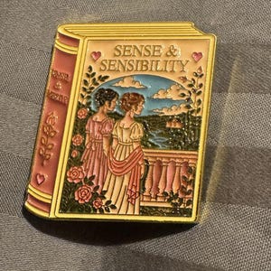May include: A rectangular enamel pin shaped like a book with a pink spine and cream cover. The front features the text SENSE & SENSIBILITY, two figures in period clothing, pink roses, and a landscape scene with a blue sky and a stone balcony.