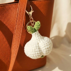 May include: A white crocheted garlic bulb keychain with green leaves attached to a gold-toned metal lobster clasp and ring, hanging from a textured orange leather tote bag. The garlic is crafted with visible yarn stitches and a rounded shape.