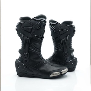 Puede incluir: Un par de botas de moto negras con diseño de caña alta y placas protectoras reforzadas en espinillas, tobillos y talones. Las botas incluyen secciones de cuero perforado en la puntera y el empeine, con detalles metálicos en la punta.