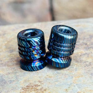 May include: Two cylindrical metal beads featuring a vibrant, iridescent finish with swirling patterns of deep blue, purple, and copper tones. Each bead has a central hole and horizontal grooved lines etched around the exterior surface.