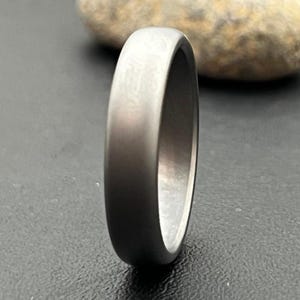 May include: A close-up of a brushed metal ring with a smooth, rounded exterior. The ring has a dark gray color and a polished interior. The ring is likely made of titanium or a similar metal. The ring is a wedding band or fashion accessory.