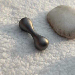 May include: A small, metallic gray dumbbell-shaped object with rounded ends and a curved center. The object is smooth and reflective, with a subtle sheen. A smooth, light-colored stone is in the background.