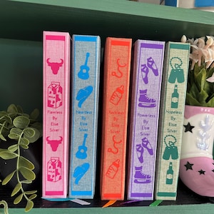 Può includere: Una collezione di sei libri con dorsi colorati, ognuno con un'icona unica. I libri sono disposti su uno scaffale con un vaso decorativo a forma di stivale pieno di fiori. I dorsi dei libri sono rosa, blu, arancioni, viola e verdi.