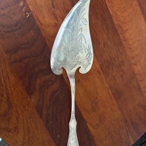 May include: A silver-toned metal cake server with a pointed, asymmetrical blade. The surface of the blade features an intricate engraving of a bird standing in tall grass near water. The handle has a slender, decorative design with etched details.