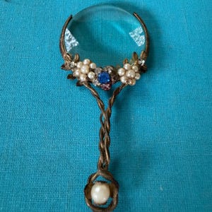 May include: A handheld magnifying glass with a circular lens framed in antiqued bronze metal. The handle features a twisted design with a single pearl at the base. The frame is decorated with clusters of white pearls and a central blue gemstone.