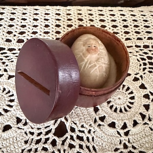May include: A round, dark purple wooden box with a slot on the lid, containing a small, off-white, egg-shaped object. The object features a carved, painted face of a person with light skin, brown hair, and subtle facial features, nestled inside.