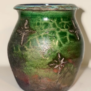 Puede incluir: Un jarrón de cerámica hecho a mano con un cuerpo redondeado y un borde ligeramente ensanchado. El jarrón presenta un vibrante esmalte verde con diseños plateados en forma de estrella. La base hace la transición a un tono más oscuro y terroso, creando un contraste llamativo. El borde es azul oscuro.