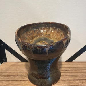 May include: A ceramic bowl with a pedestal base, featuring a glossy glaze in shades of dark brown, tan, and cream. The interior of the bowl displays a swirling, mottled pattern of light and dark earth tones, while the exterior has a smooth, dark brown finish that tapers inward toward the center before widening at the base.