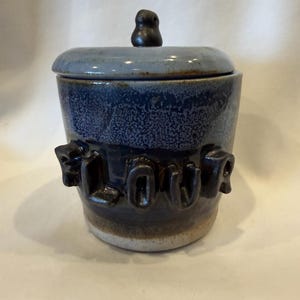 Può includere: Un contenitore per farina in ceramica fatto a mano con coperchio. Il contenitore è blu scuro con una trama maculata e la parola "FLOUR" è in rilievo sul lato. Il coperchio è blu chiaro con un piccolo pomello scuro.
