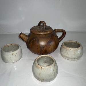 Puede incluir: Un juego de té de cerámica hecho a mano con una tetera marrón con tapa y asa, acompañado de tres tazas de té pequeñas, redondas y de color claro. El juego tiene una estética rústica y terrosa.