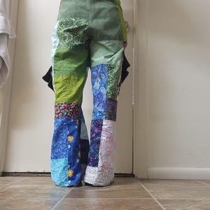 May include: Patchwork pants featuring a variety of fabric squares in shades of green, blue, purple, and floral patterns. The trousers have a flared leg silhouette, an elasticized waistband, and black side pockets visible on the hips.