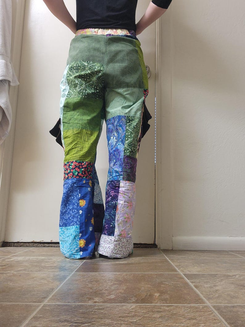 May include: Patchwork pants featuring a variety of fabric squares in shades of green, blue, purple, and floral patterns. The trousers have a flared leg silhouette, an elasticized waistband, and black side pockets visible on the hips.