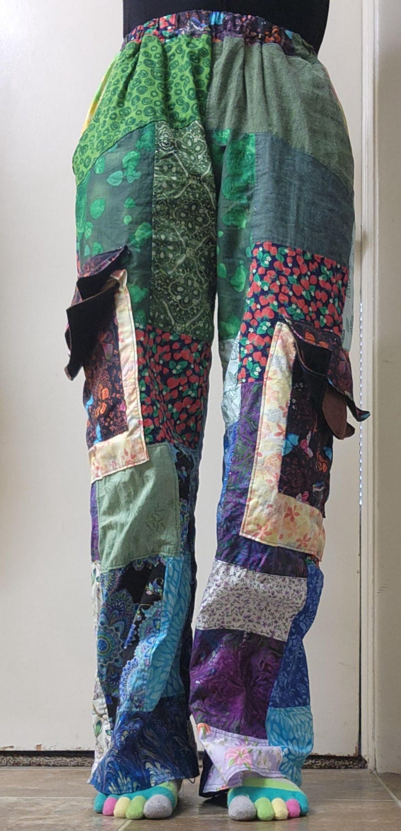 May include: Patchwork pants featuring a variety of fabric squares in shades of green, blue, purple, and red with floral, paisley, and geometric patterns. The pants have an elastic waistband and two large cargo-style pockets on the outer thighs.